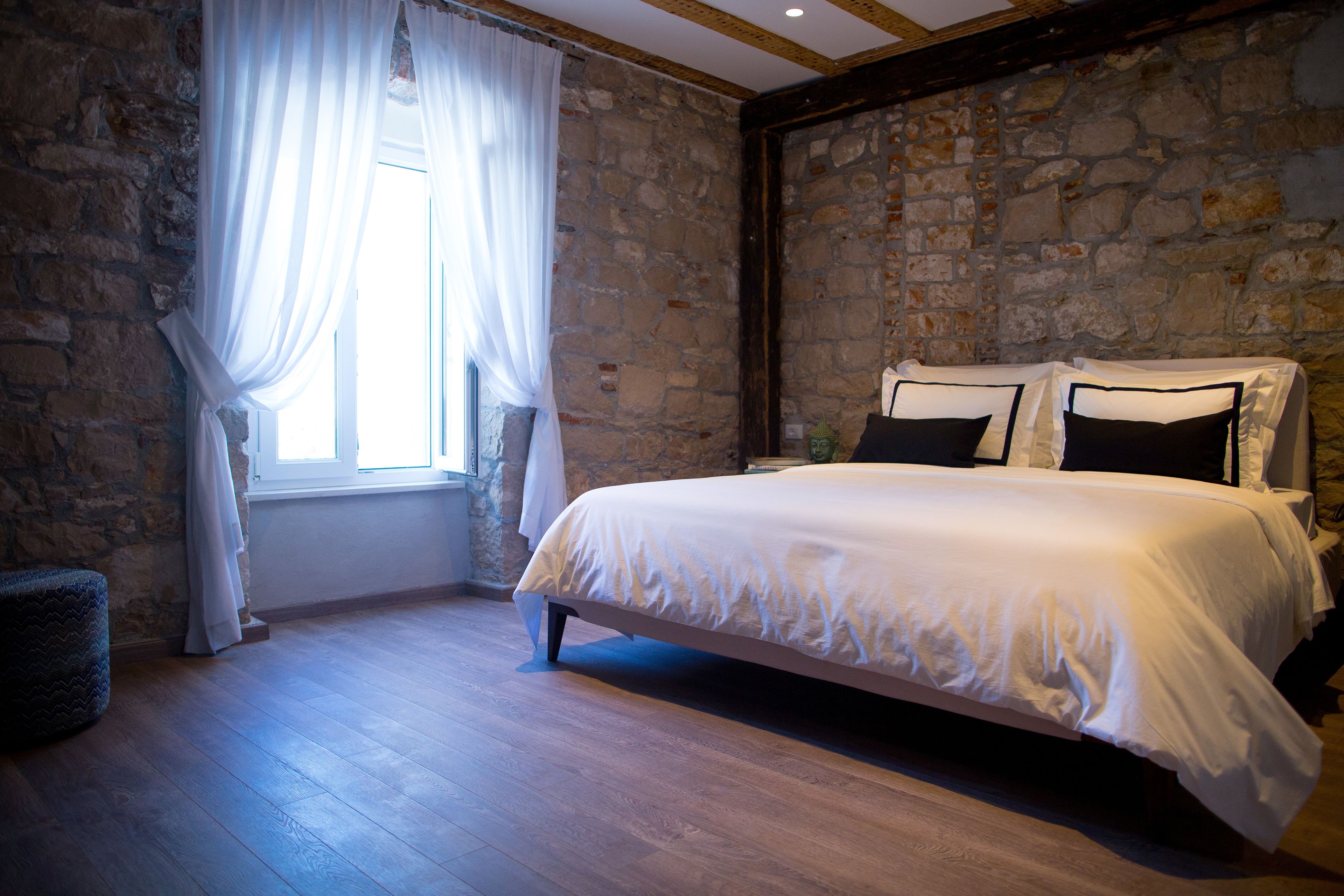 Ein Schlafzimmer mit einer Steinwand, einem großen Bett mit weißer Bettwäsche und einem Fenster mit transparenten Vorhängen.