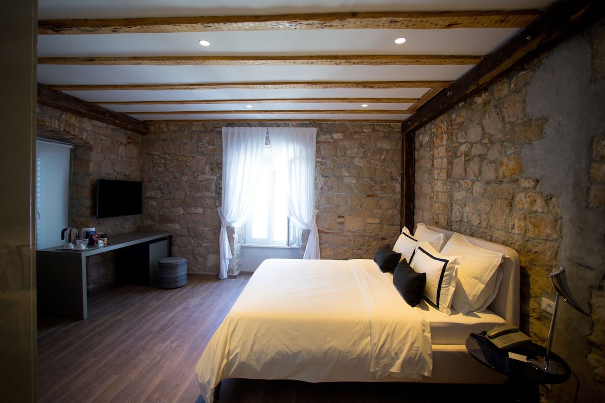 Un dormitorio con pared de piedra, vigas de madera, cama con ropa blanca, mesita de noche, lámpara y televisor sobre un mueble.
