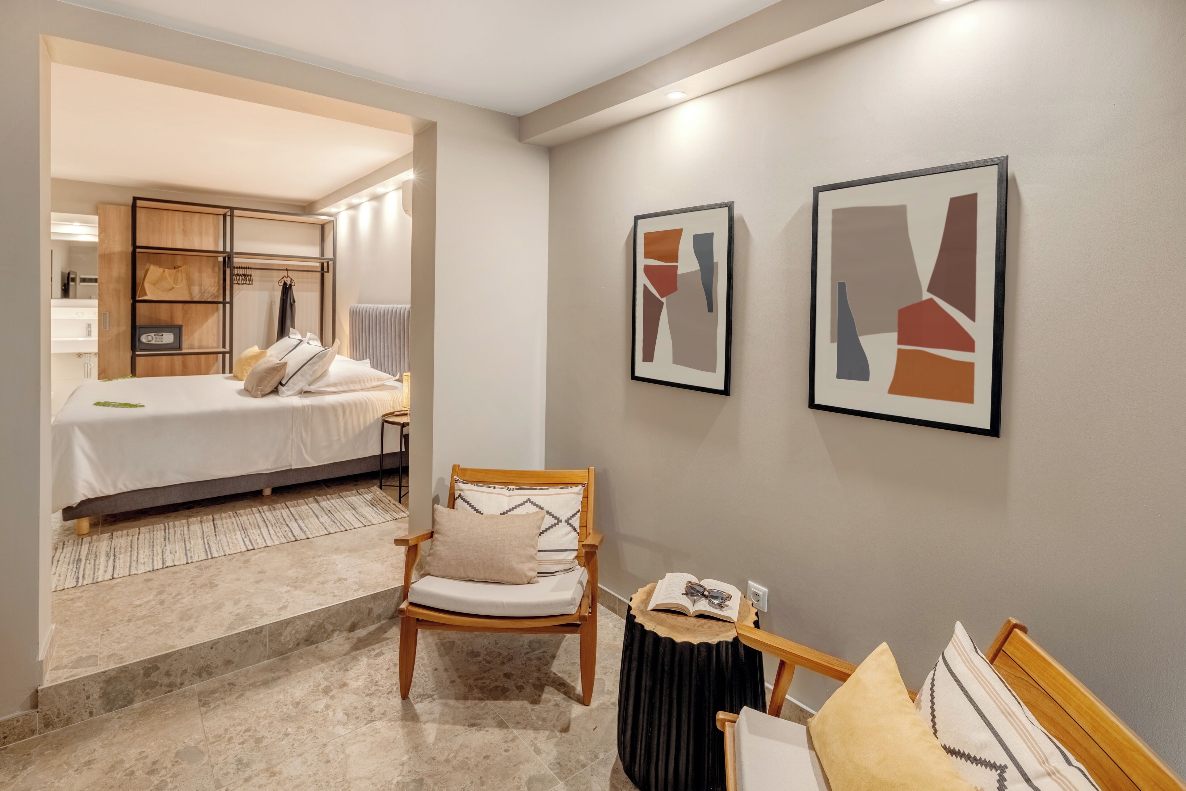 A hotel room with a bed, a chair, a small table with a book, and abstract art on the wall.