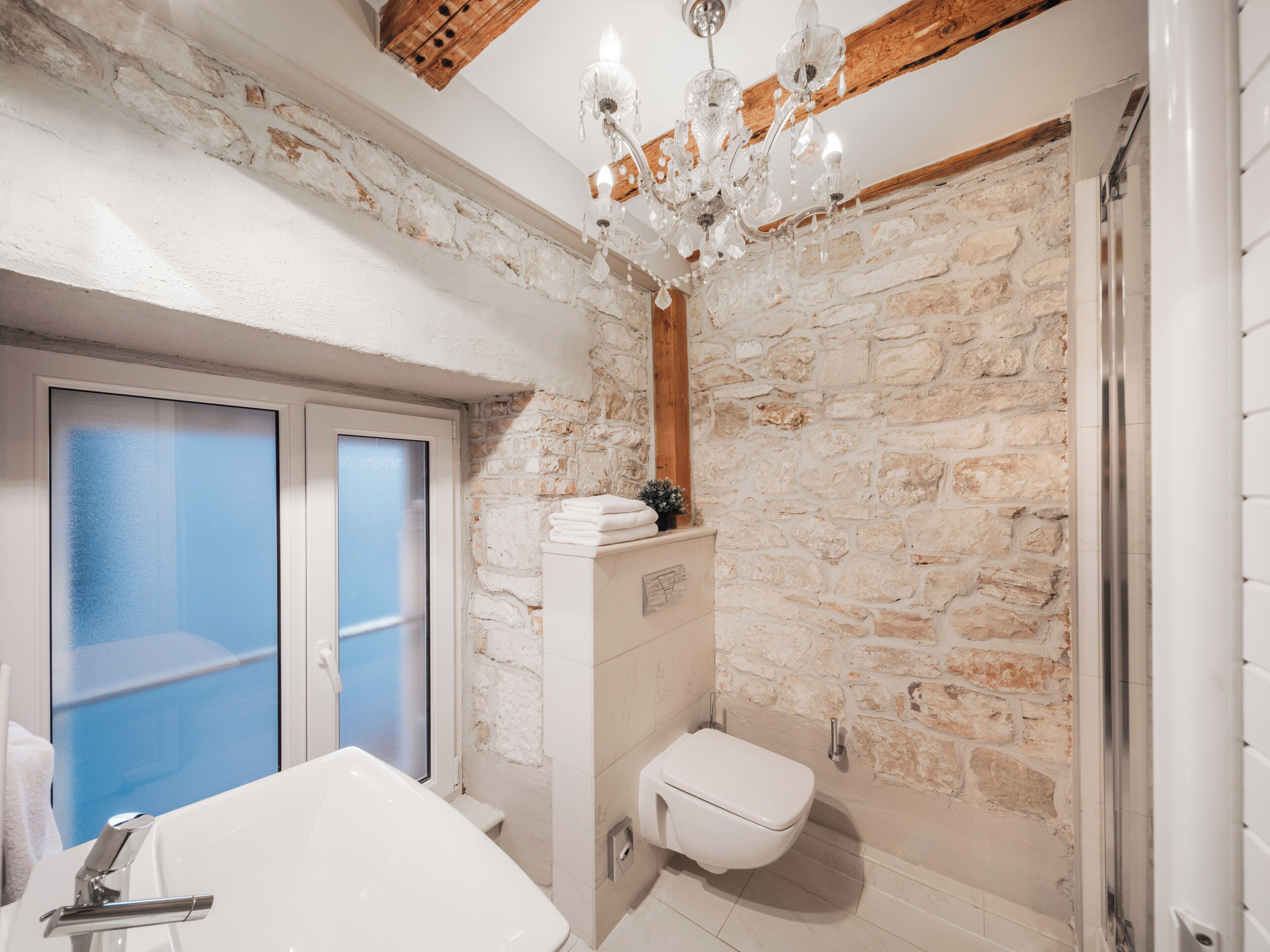 A bathroom with a stone wall, a white bathtub, a white toilet, and a chandelier.