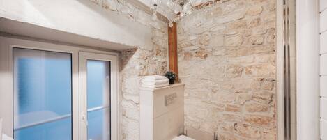 A bathroom with a stone wall, a white bathtub, a white toilet, and a chandelier.