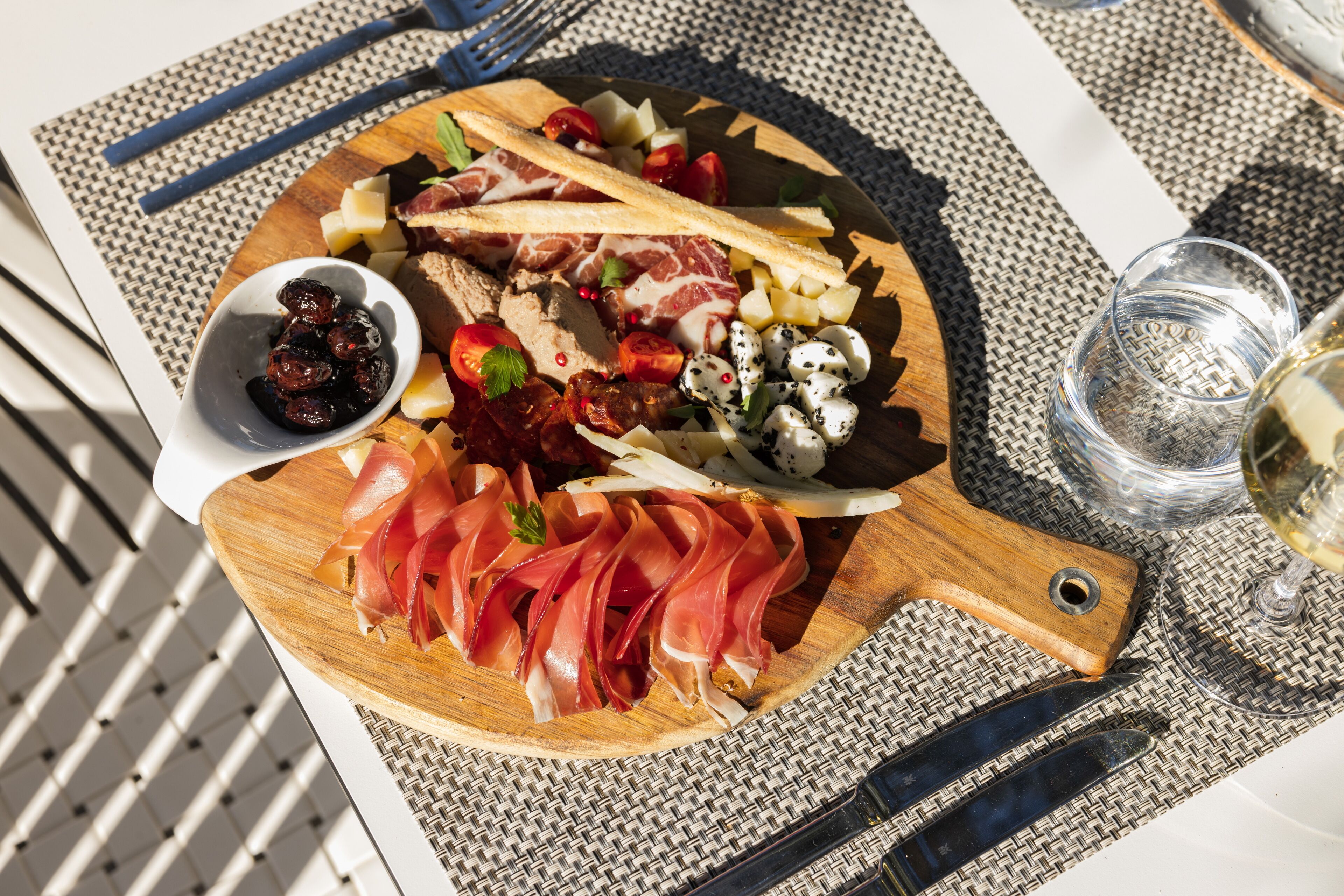 A wooden board with various meats, cheeses, and olives on a woven table mat.