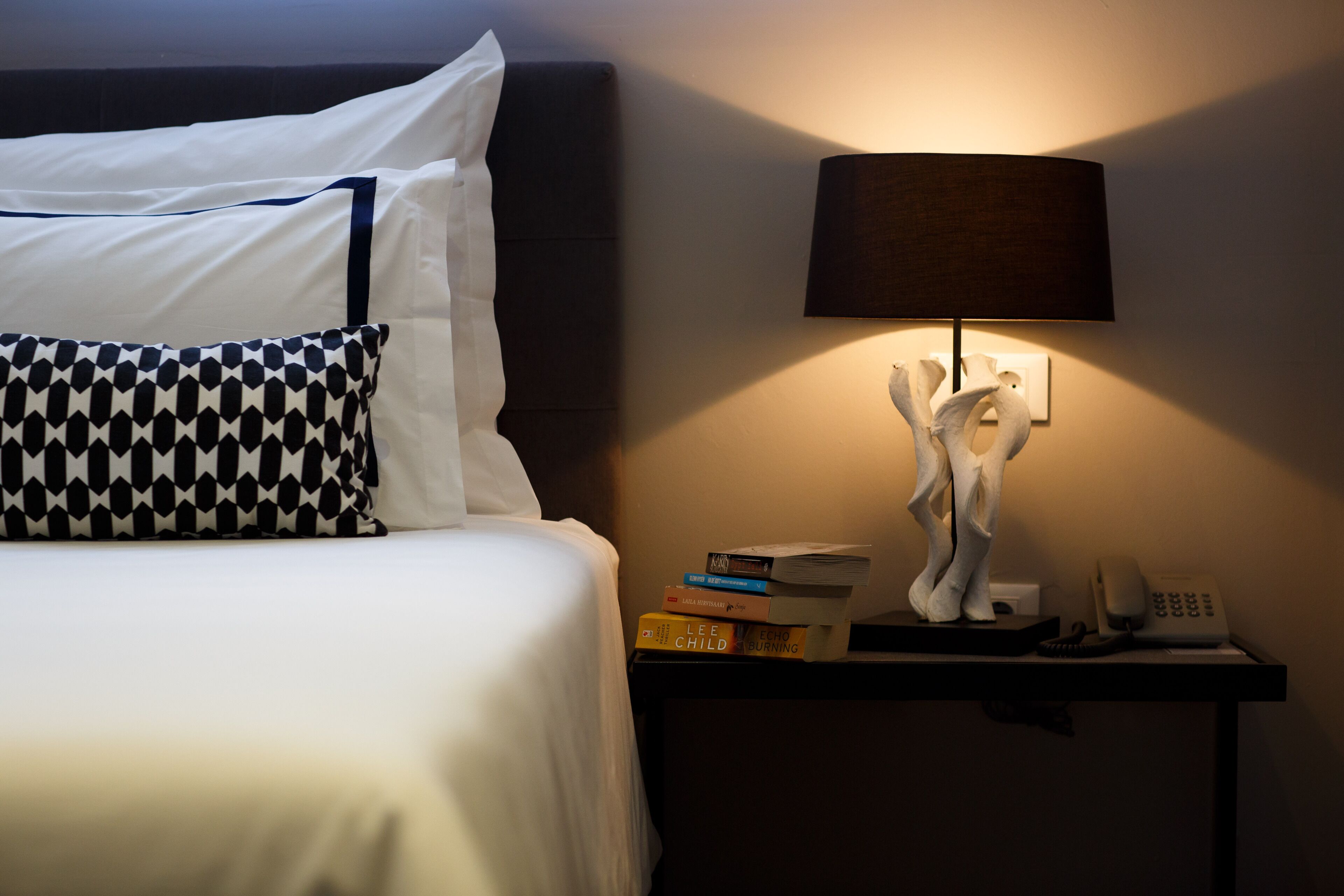 A neatly made bed with white and black patterned pillows, a bedside table with a lamp, books, and a phone.