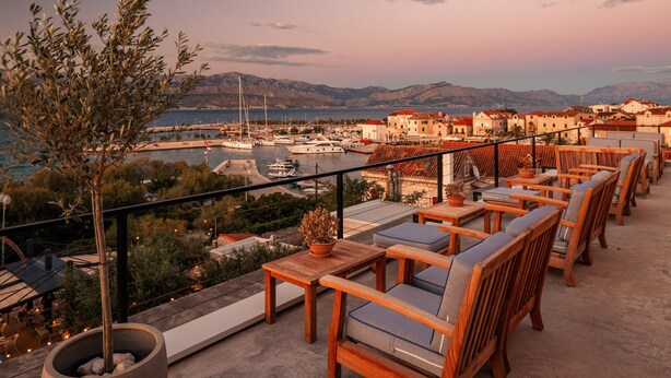 A terrace with wooden furniture, a potted plant, and a view of a marina and buildings in the distance.