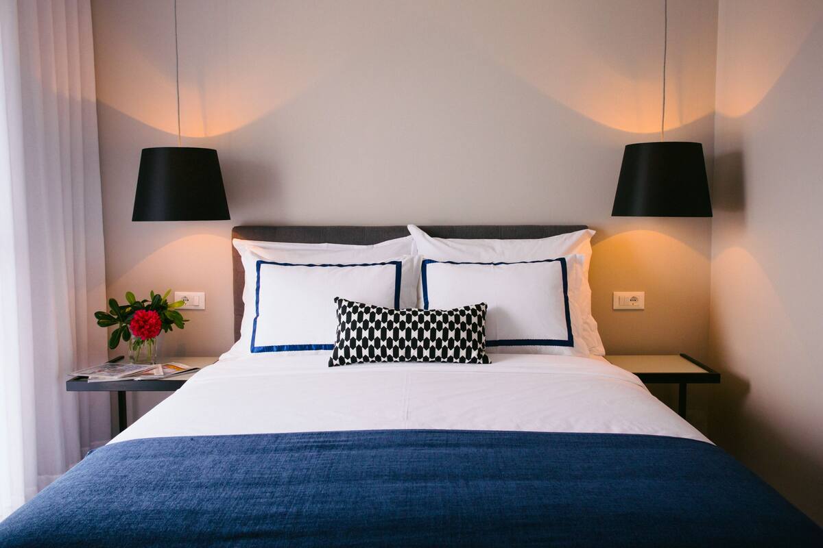 A neatly made bed with white and blue bedding, two black lamps on either side, and a small table with a vase of flowers.
