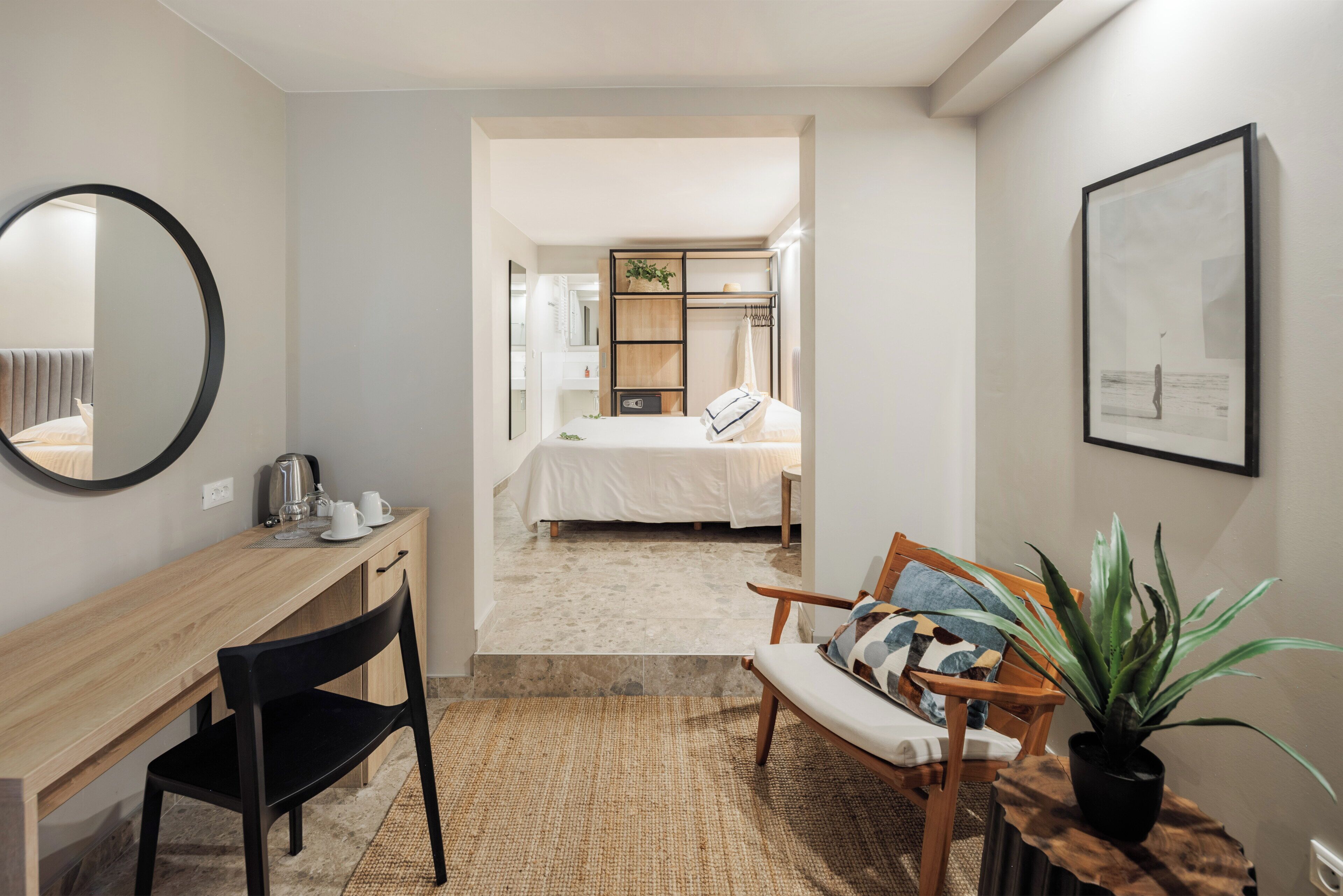 Una habitación de hotel moderna con un escritorio de madera, un sillón cómodo, una cama con edredón blanco, una mesita con una planta y un cuadro en la pared.
