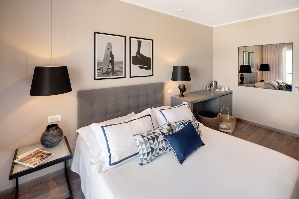 Una habitación de hotel moderna con una cama grande, dos lámparas de noche, un cabecero tapizado color gris, una mesita de noche con un florero y una revista, y dos cuadros enmarcados en la pared.