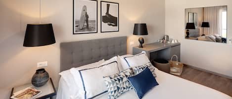 Une chambre d’hôtel moderne avec un grand lit, deux lampes de chevet, une tête de lit rembourrée de couleur grise, une petite table de chevet avec un vase et un magazine, et deux tableaux encadrés accrochés au mur.