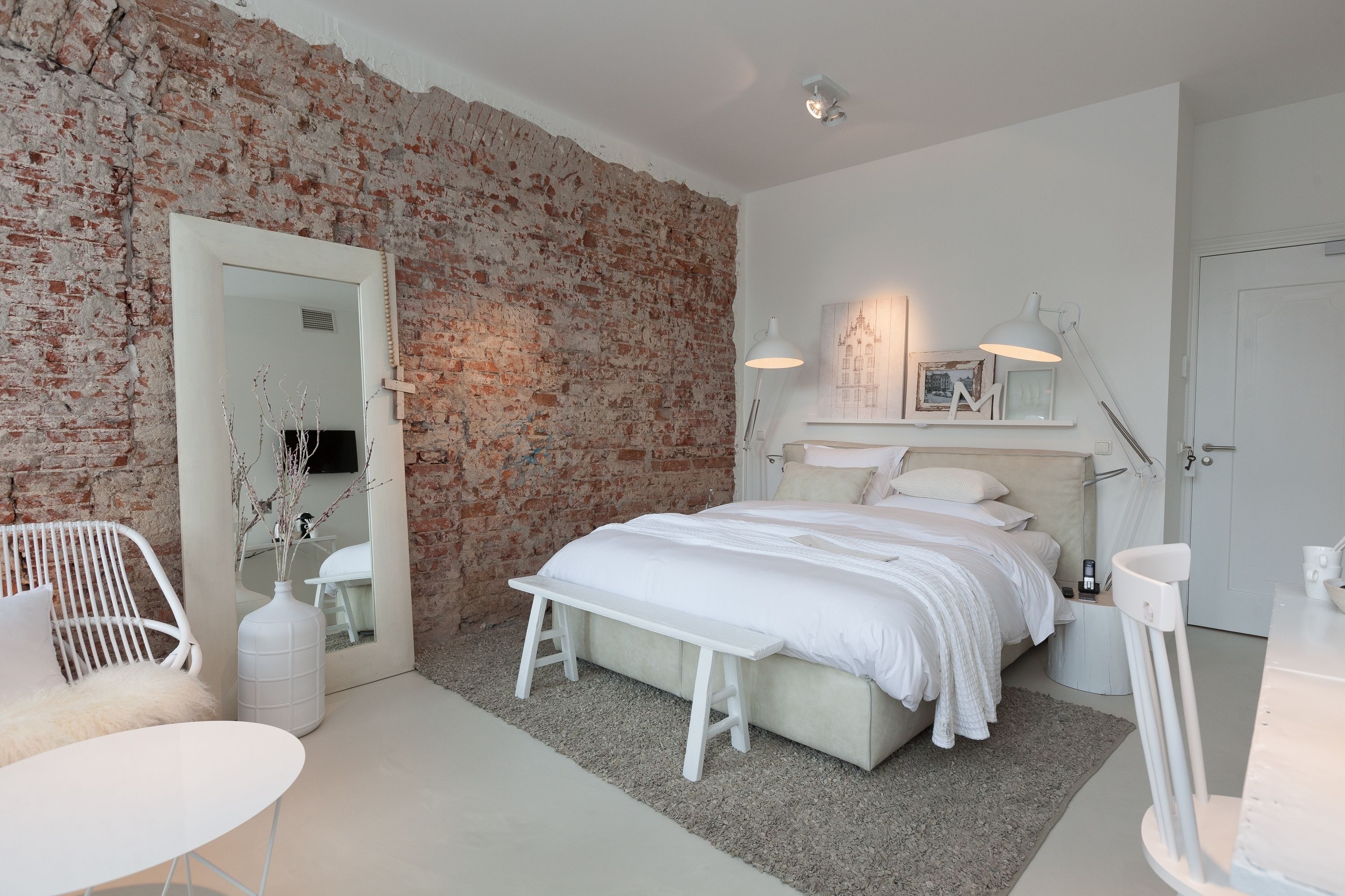 A bedroom with a brick wall, a bed with white bedding, a bedside table, a lamp, and a chair.