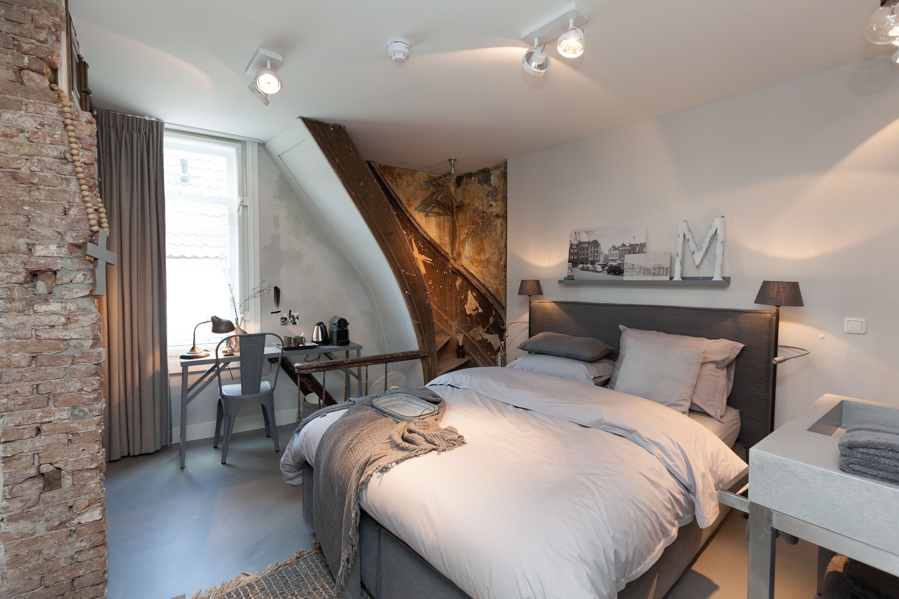 Une chambre moderne avec un plafond en pente, un lit doté d’une tête de lit grise, un bureau avec une chaise et une grande fenêtre avec des rideaux.