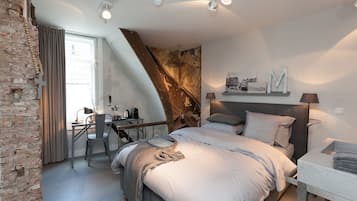 Une chambre moderne avec un plafond en pente, un lit doté d’une tête de lit grise, un bureau avec une chaise et une grande fenêtre avec des rideaux.