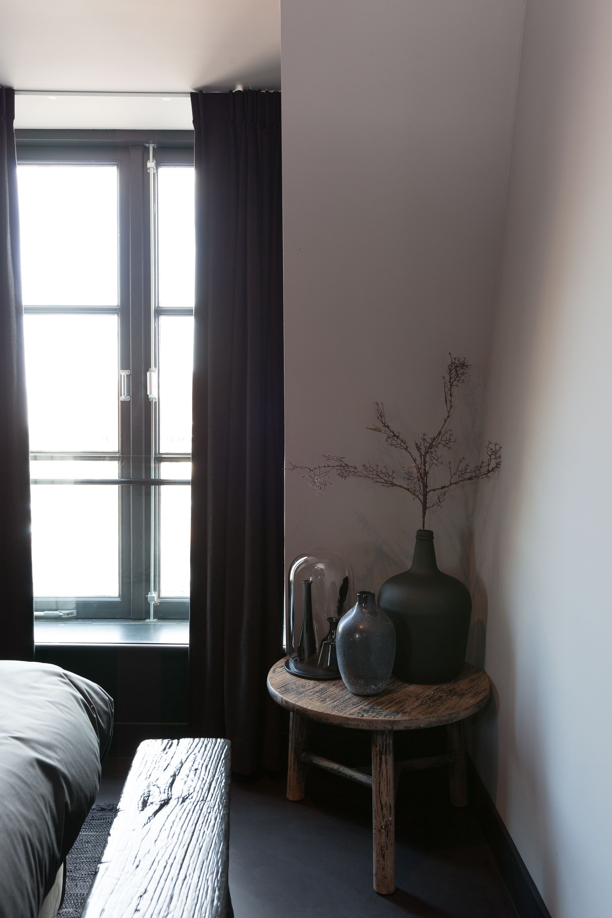 A bedroom with a bed, a window with curtains, a bedside table with a vase, and a wall-mounted shelf.