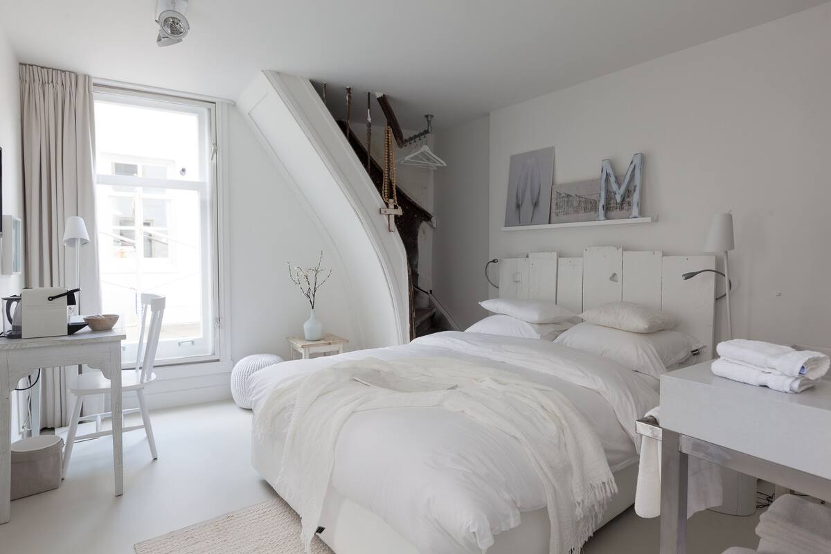 A bedroom with a large bed, a desk, and a chair. There is a window with curtains, a painting on the wall, and a coat rack.