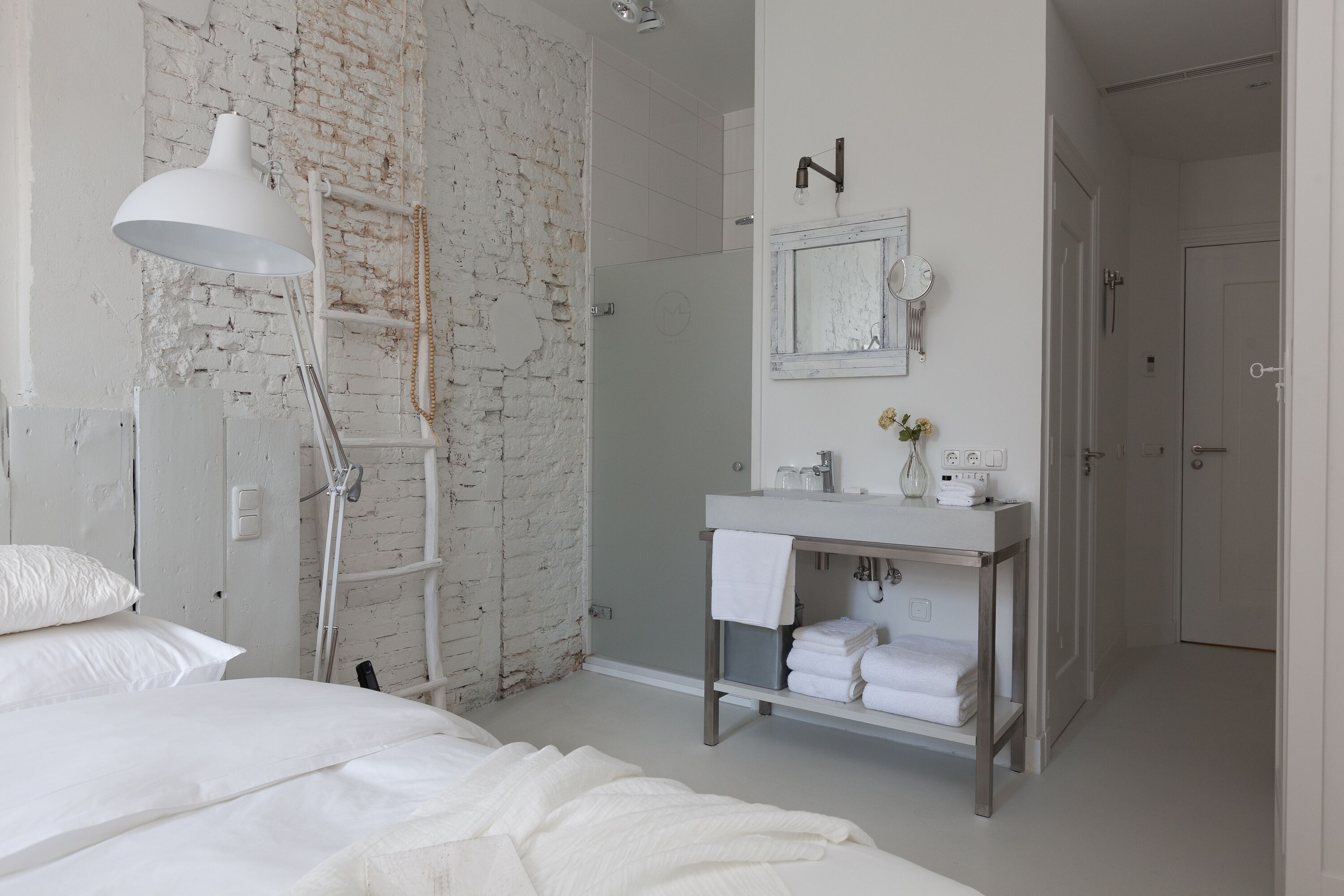Une chambre à coucher avec un lit, une lampe blanche, un mur en briques, une salle de bain avec un lavabo et un miroir, et une porte.
