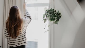A person adjusting a window blind in a room with a white interior, a hanging plant, and a staircase.