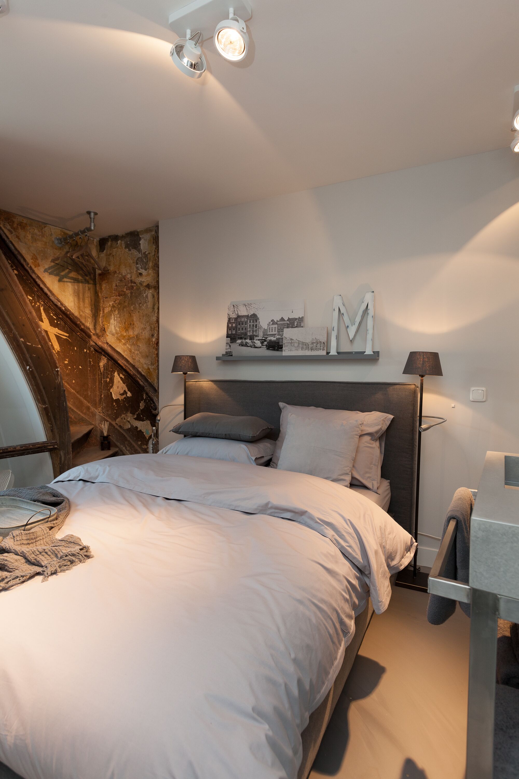 A modern bedroom with a large bed, a headboard, a nightstand, a lamp, and a wall-mounted shelf with a picture and a letter 'M'.