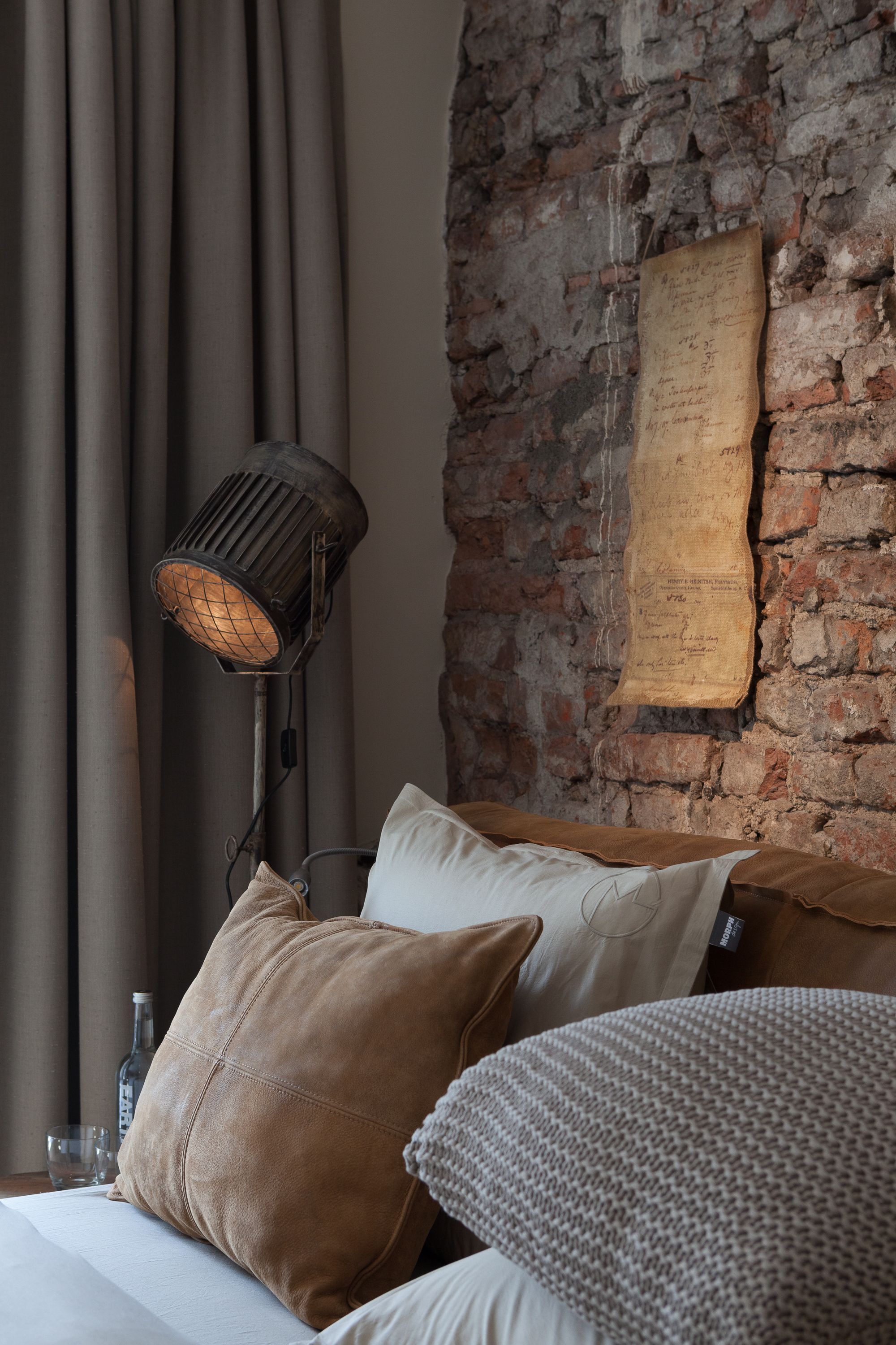 A room with a brick wall, a hanging parchment, a floor lamp, and several pillows.