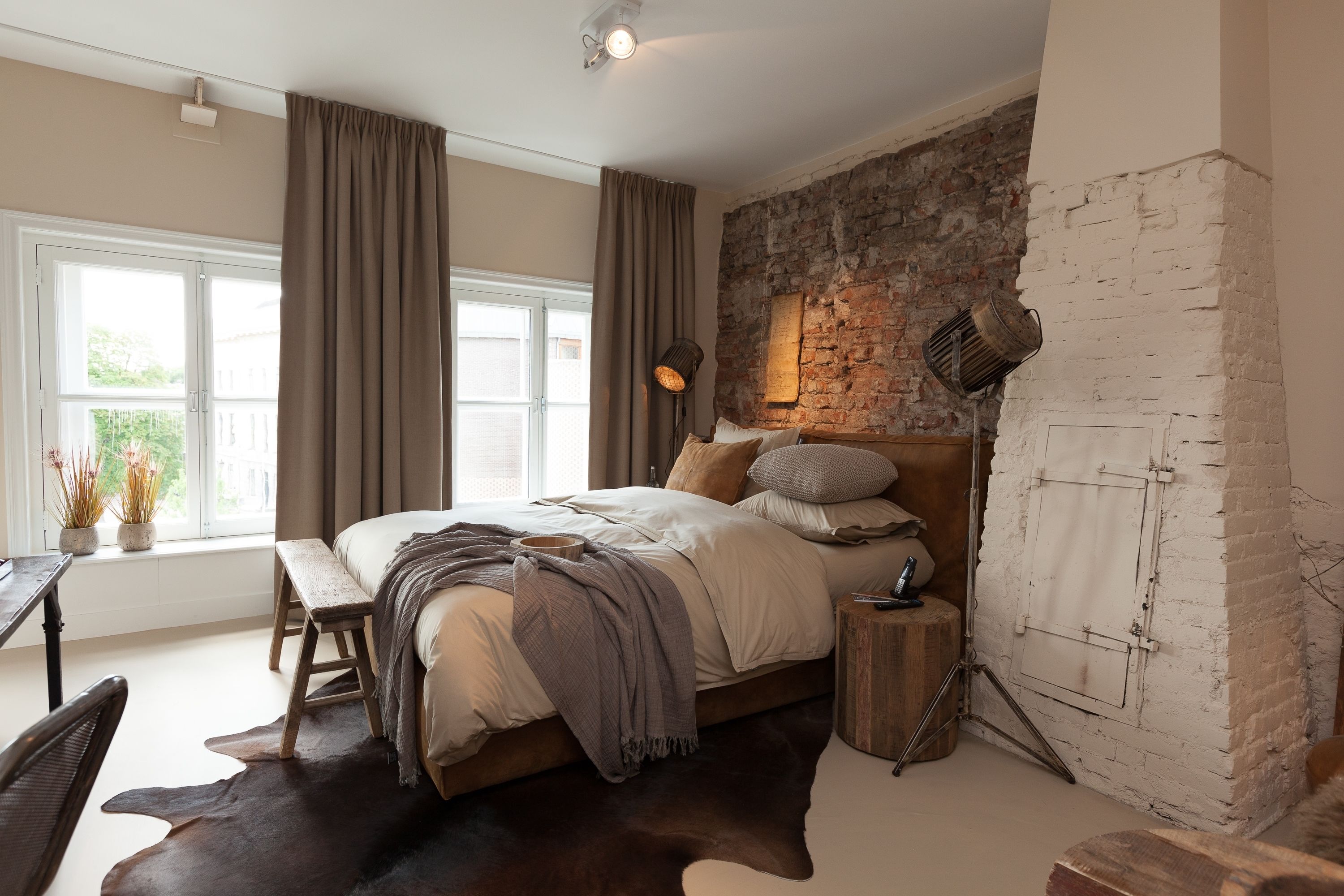 A bedroom with a large bed, a wooden bench, a rustic brick wall, and a window with curtains.