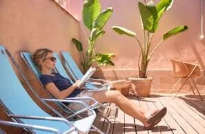 A person relaxing on a sun lounger with a book, surrounded by potted plants and a tiled wall.