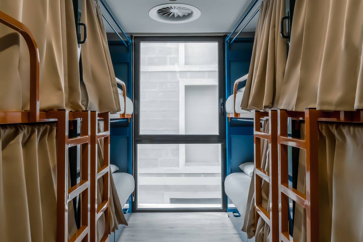 A narrow corridor with bunk beds on either side, a window at the end, and a ceiling light.