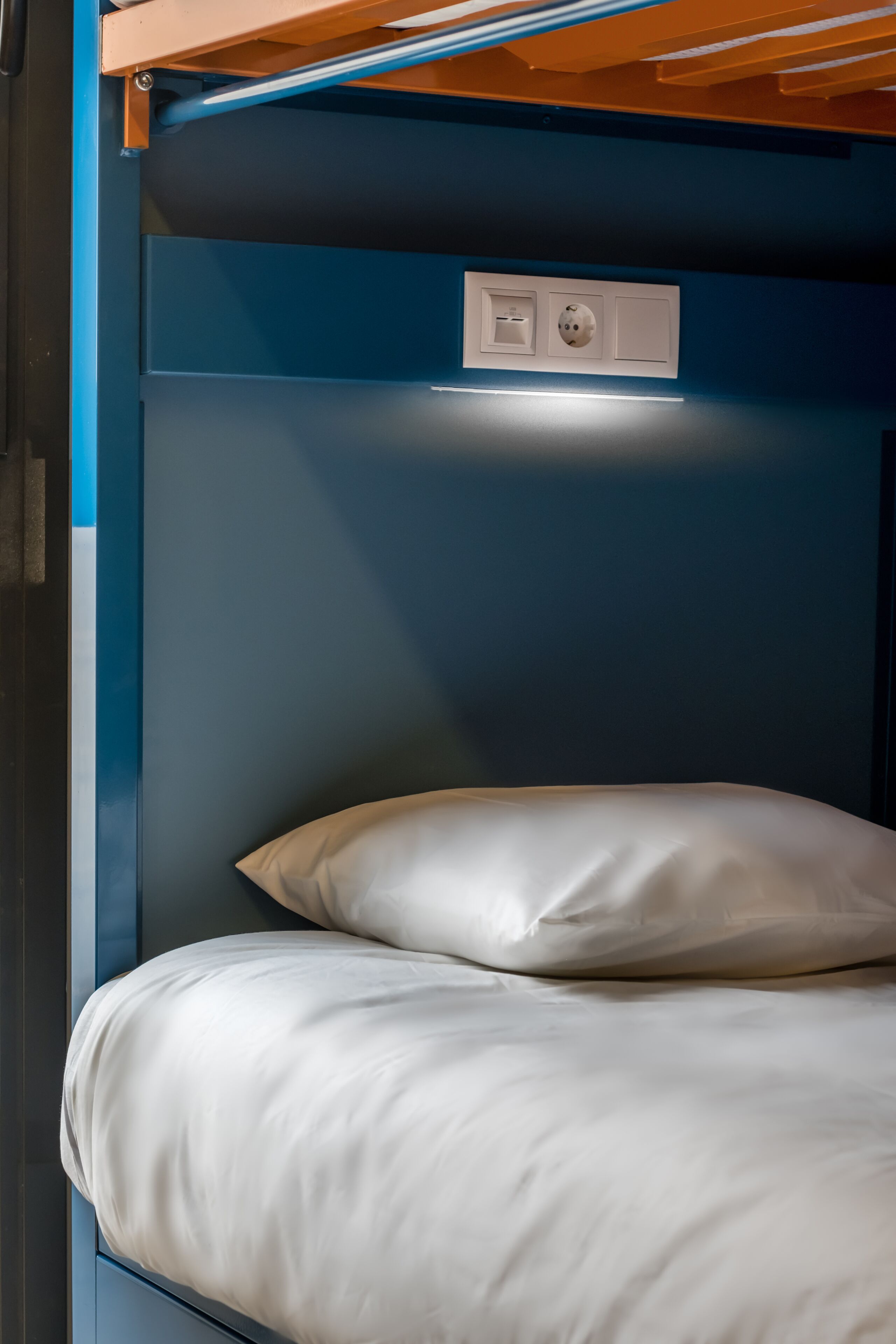 A bunk bed with a white mattress and pillow, a blue wall, and a power outlet above the bed.