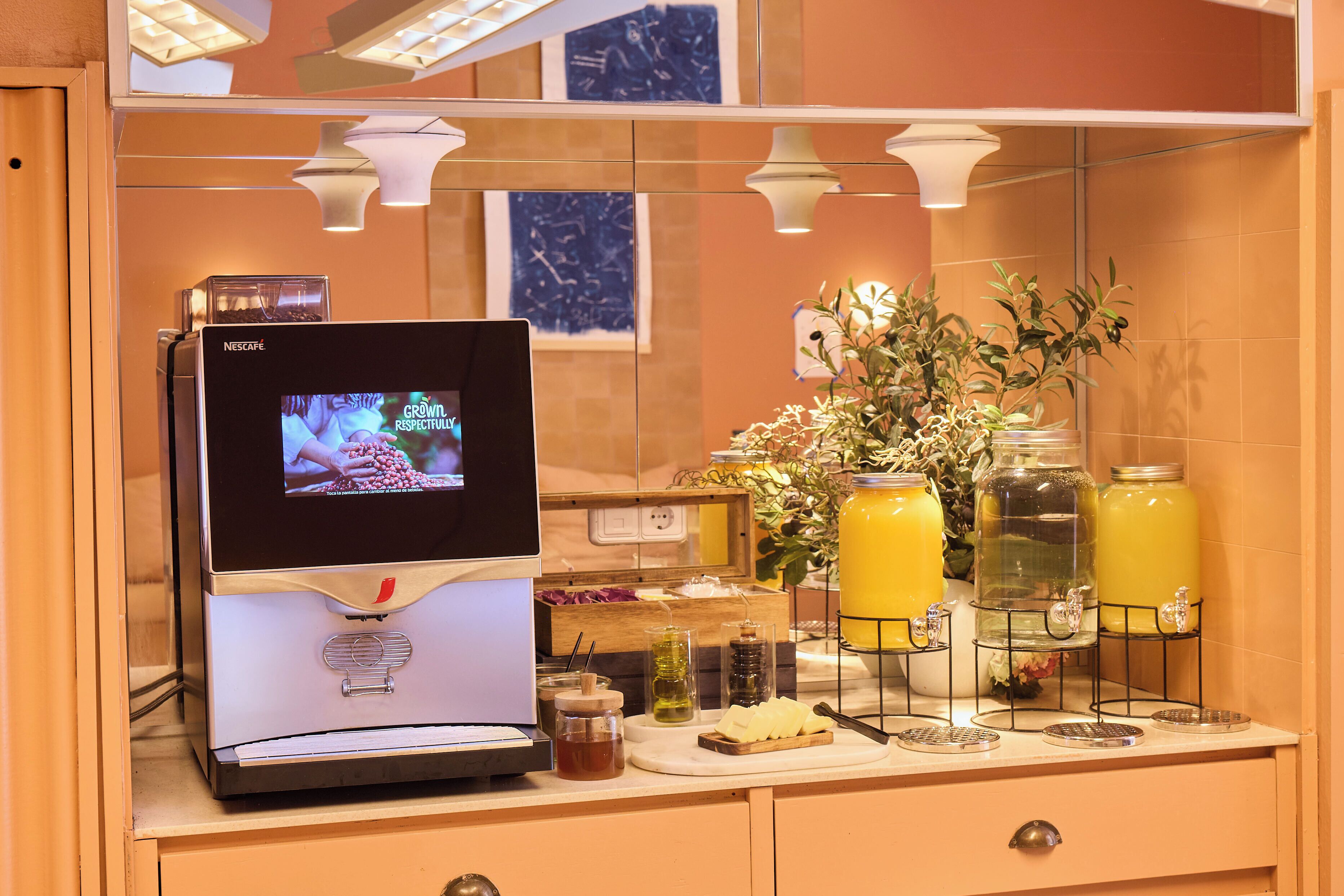 A Nescafé coffee machine with a screen, a counter with jars of honey and jam, and a potted plant.