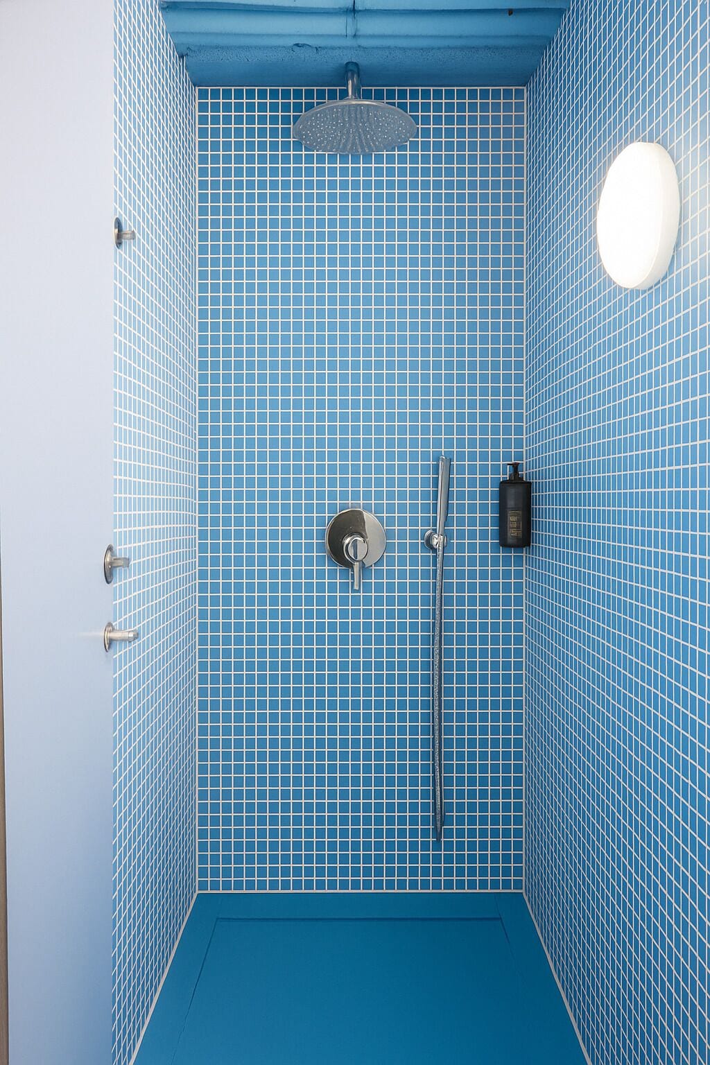 Un baño pequeño con azulejos, suelo y paredes azules, un cabezal de ducha manual, un grifo empotrado y un dispensador de jabón.