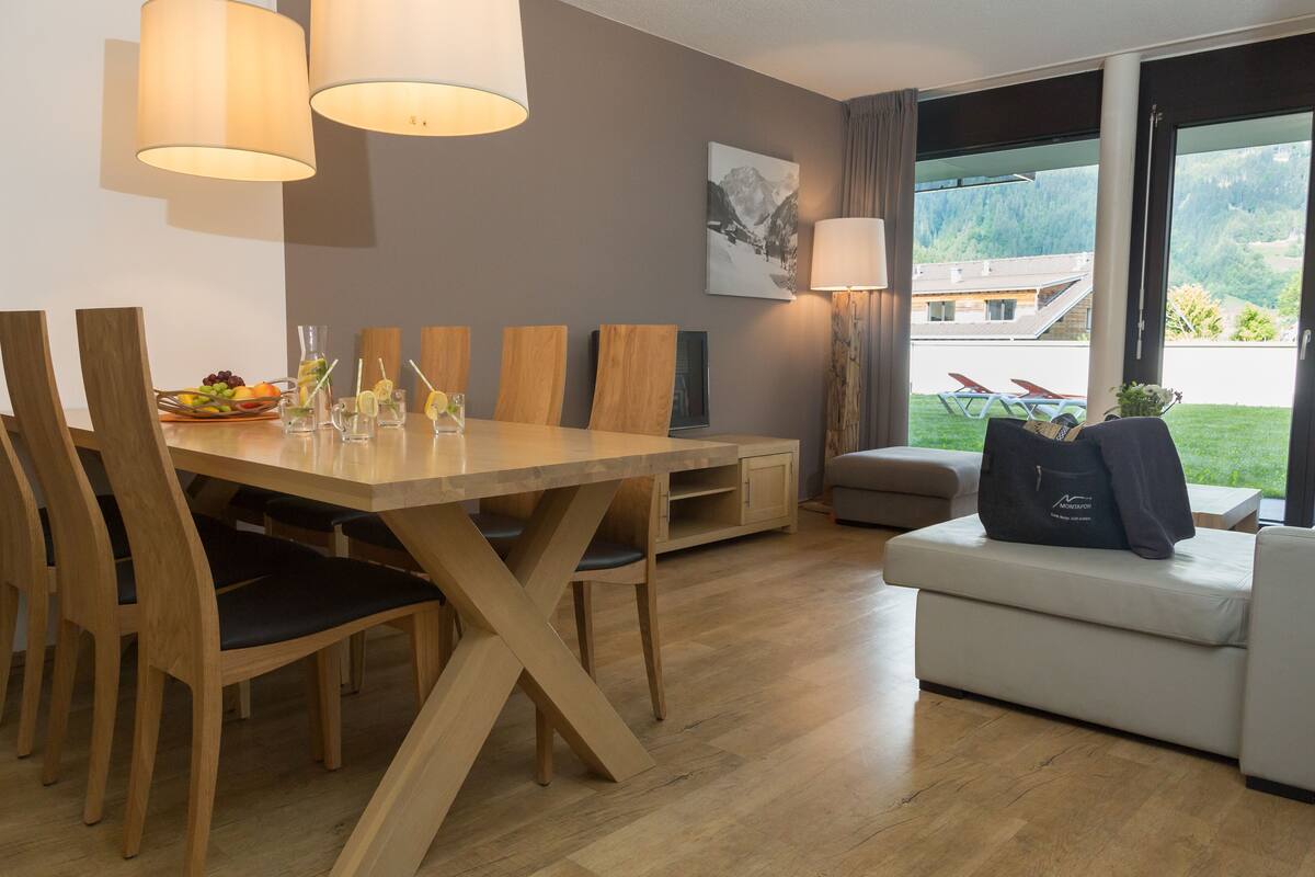 A modern dining area with a wooden table and chairs, a sofa, and a view of a mountainous landscape through large windows.