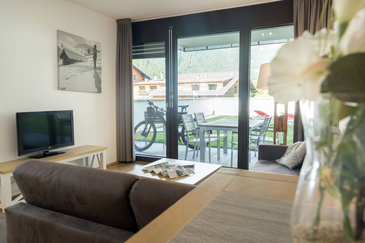 A modern living room with a sofa, a coffee table with magazines, a TV, and a large window with a view of a mountain and a building.