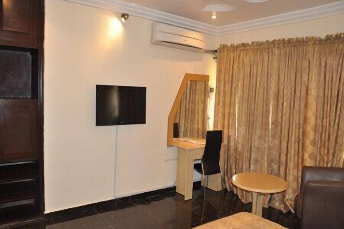 Une chambre d’hôtel équipée d’un téléviseur à écran plat fixé au mur, d’un bureau en bois, d’une chaise, d’une petite table ronde et d’une grande fenêtre avec des rideaux.