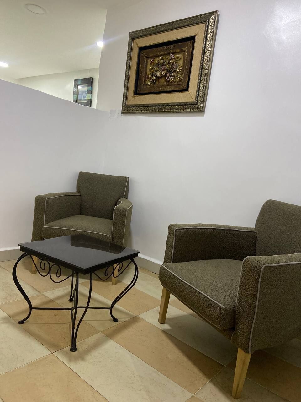 A room with two armchairs and a small table, a framed artwork on the wall, and a staircase in the background.