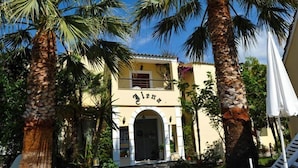 A building with a yellow facade, an arched entrance, and palm trees in the foreground. - Ilena Hotel (Corfu)