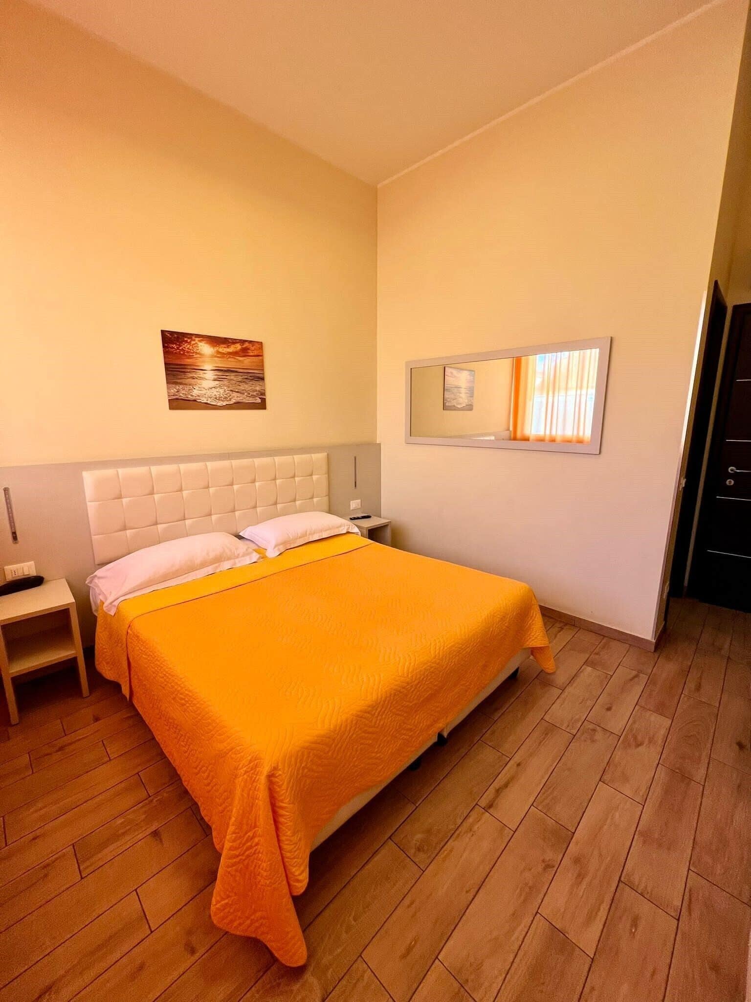 A bedroom with a large bed, a nightstand, a bedside table, a wall-mounted mirror, and a painting on the wall.