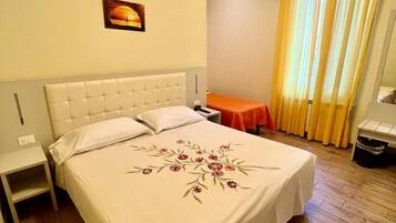 A hotel room with a large bed, a nightstand, a small table, a chair, a window with curtains, and a wall-mounted picture.