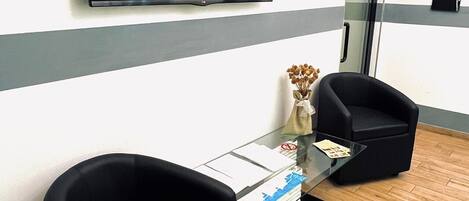 Un espace d’attente moderne, doté d’un téléviseur à écran plat fixé au mur, de deux fauteuils noirs, d’une table basse en verre sur laquelle sont posés des documents, et d’un plancher en bois.