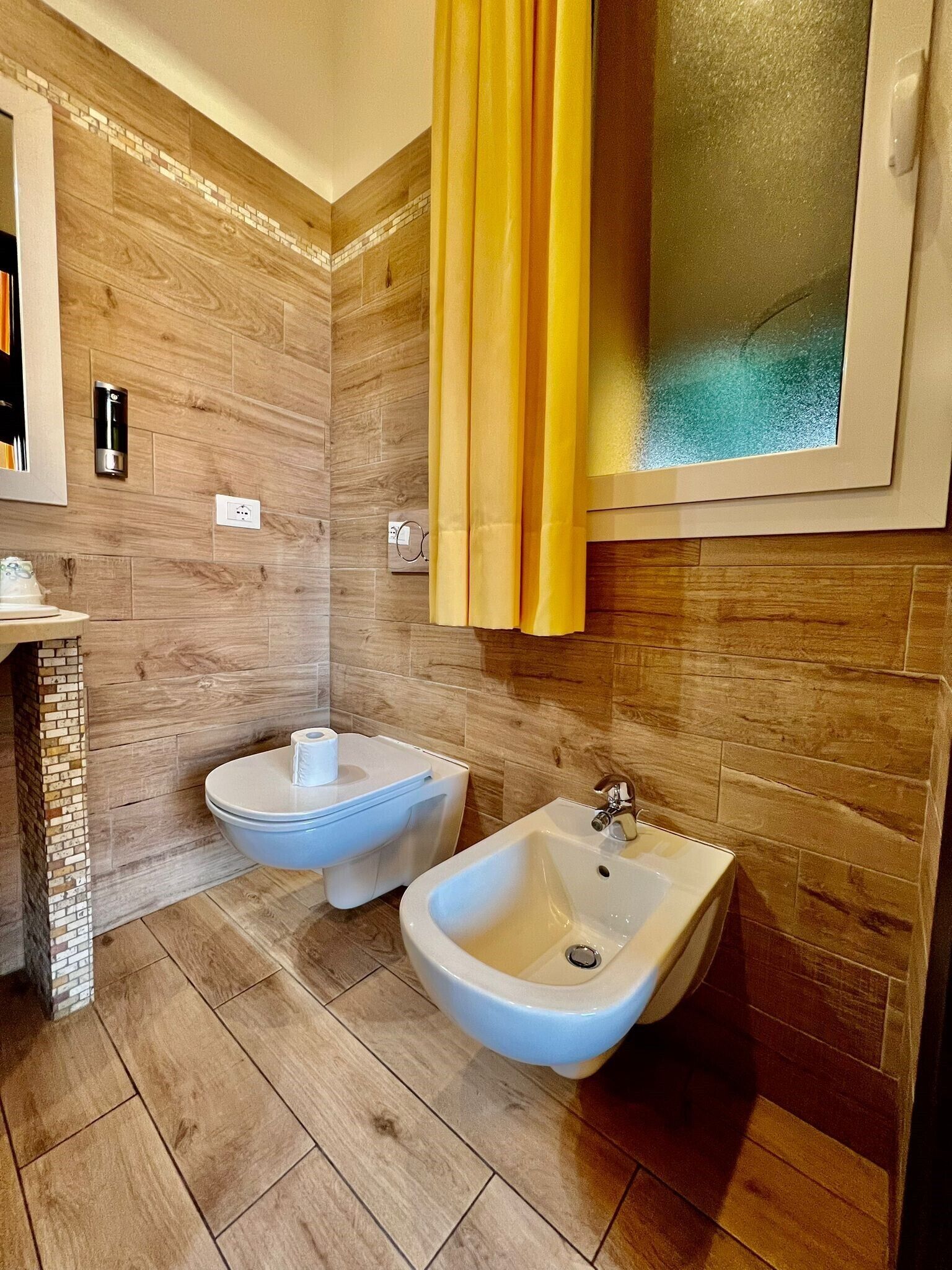 Un bany modern amb terra de fusta, dos bidet blancs, un mirall i una cortina groga.