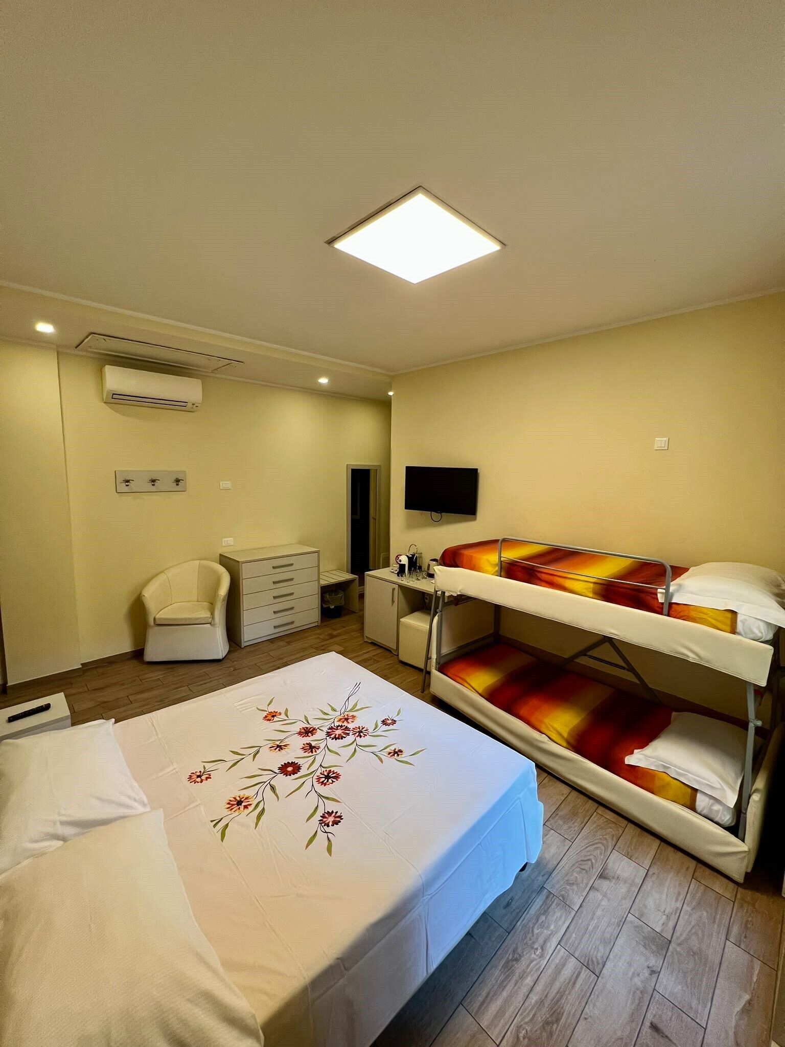 A hotel room with a bunk bed, a desk, a chair, a flat-screen TV, an air conditioning unit, and a ceiling light.