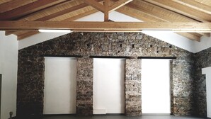 Una habitación amplia con techo alto y vigas de madera a la vista. Cuenta con una pared de piedra y tres puertas blancas grandes.
