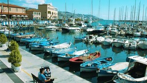Un puerto deportivo con numerosas embarcaciones amarradas, un banco con gente sentada y edificios al fondo.