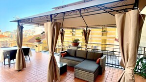 Una terraza con sofá y sillones, una mesa y una pérgola con cortinas.