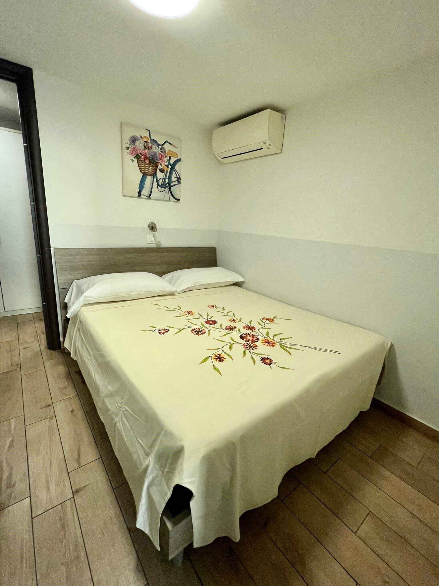 A single bed with a floral bedspread, a wall-mounted air conditioner, and a painting of a bicycle and flowers on the wall.