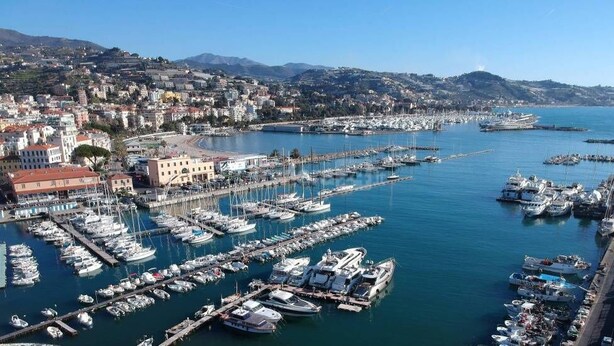 Un puerto deportivo con numerosas embarcaciones amarradas, un paseo marítimo y un fondo con una zona urbana densamente poblada y colinas.