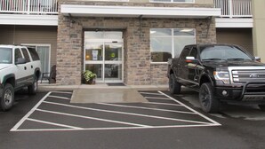 Un edificio de varios pisos con fachada de piedra, una entrada central con puertas de vidrio y un estacionamiento con dos camionetas SUV.