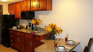 Una cocina con armarios de madera, un refrigerador negro, un microondas, encimeras de granito, un lavamanos y una zona de comedor con platos y una jarra de flores.