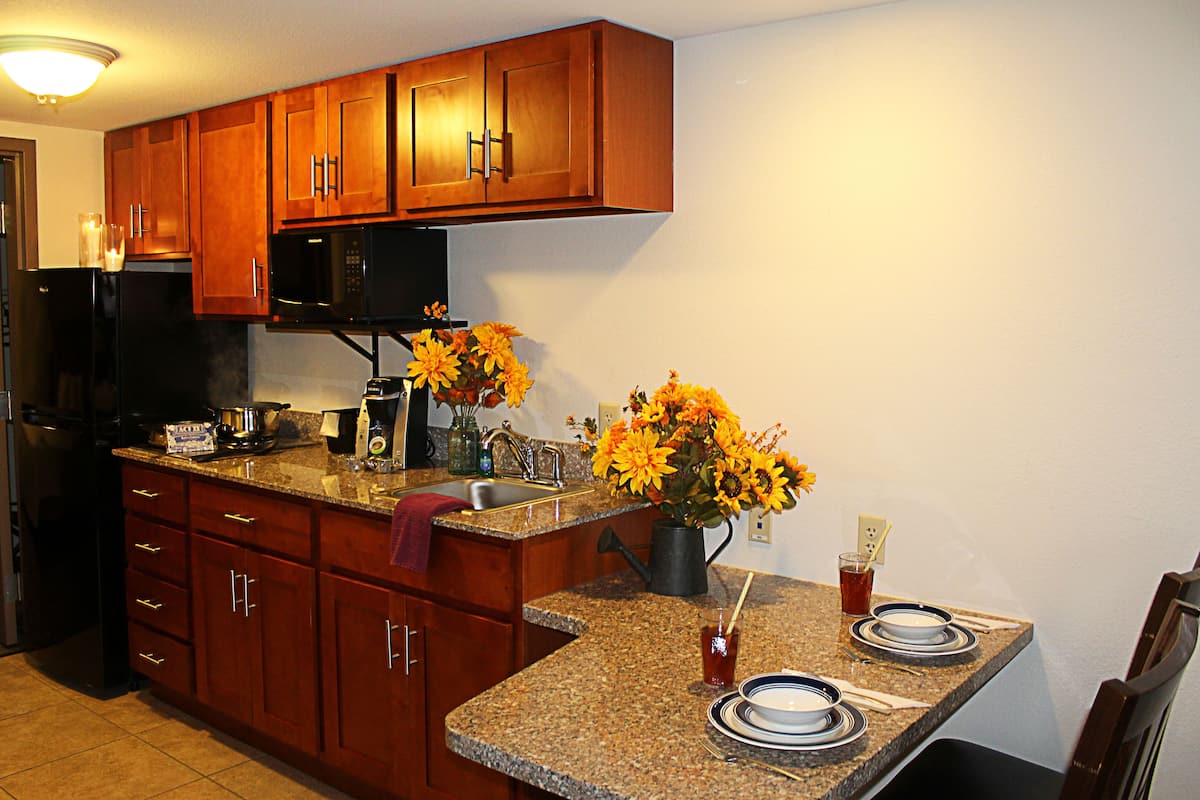 A kitchen with wooden cabinets, a black refrigerator, a microwave, granite countertops, a sink, and a dining area with plates and a pitcher of flowers.