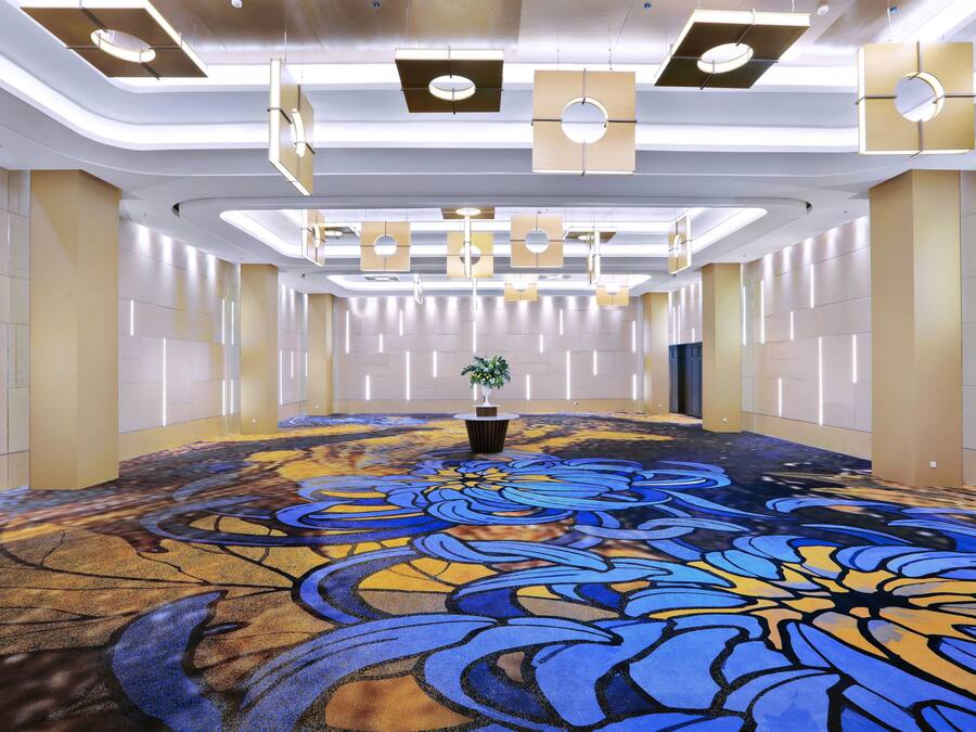 A spacious, modern hotel lobby with a large, colorful carpet, recessed lighting, and a central plant.