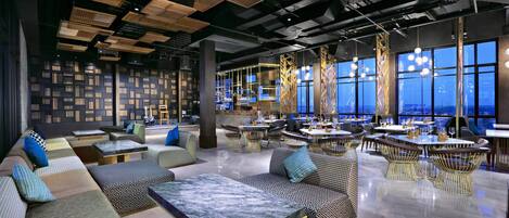 A modern lounge area with seating arrangements, tables, and a view of the cityscape through large windows.