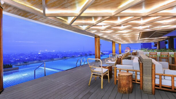A rooftop pool area with wooden deck, lounge chairs, and a view of a cityscape at dusk. - The Alts Hotel (Palembang)