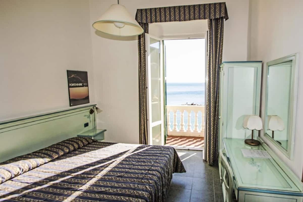 A hotel room with a bed, a dresser, a mirror, and a view of the sea through an open door.