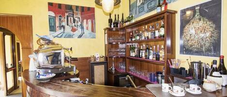A bar area with wooden shelves stocked with bottles and glasses, a vintage radio, and a large painting of a street scene.